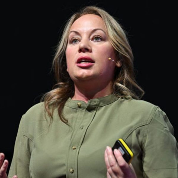 Sarah Armstrong-Smith is doing a speech. She has a microphone connected to her ear and is wearing a green shirt. She is stood against a black background and it is clear she is using hand gestures to emphasize her point.